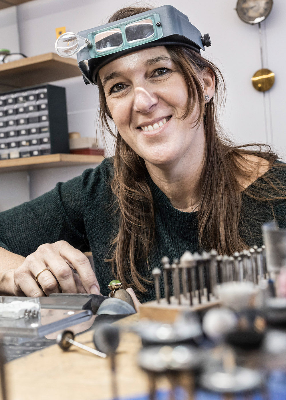 Lächelnde Frau bei der Arbeit in einer Schmuckwerkstatt. Sie trägt ein Stirnband mit Vergrößerungsglas. Auf dem Schreibtisch ist sie von Werkzeugen und Geräten zur Schmuckherstellung umgeben.