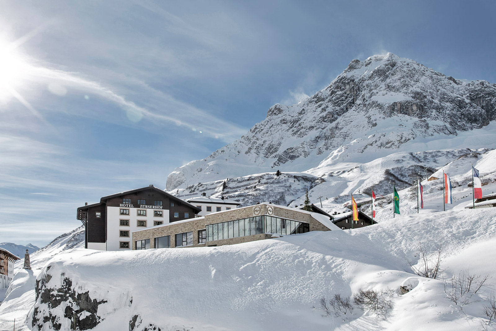 Eine verschneite Berglandschaft mit einer von Schnee umgebenen Hütte. In der Nähe des Gebäudes sind verschiedene Länderflaggen zu sehen. Eine helle Sonne scheint am klaren blauen Himmel und erhellt das majestätische Gelände.