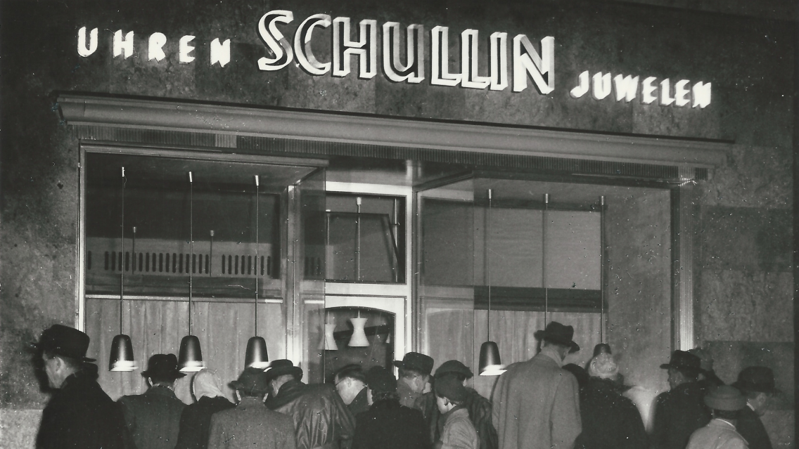 Eine Gruppe von Menschen in Hüten und Mänteln versammelt sich vor einem Juwelier- und Uhrengeschäft mit einem großen Schild, auf dem in Neonbuchstaben „UHREN SCHULLIN JUWELEN“ steht. Der Ladeneingang ist beleuchtet und bietet elegante Artikel.