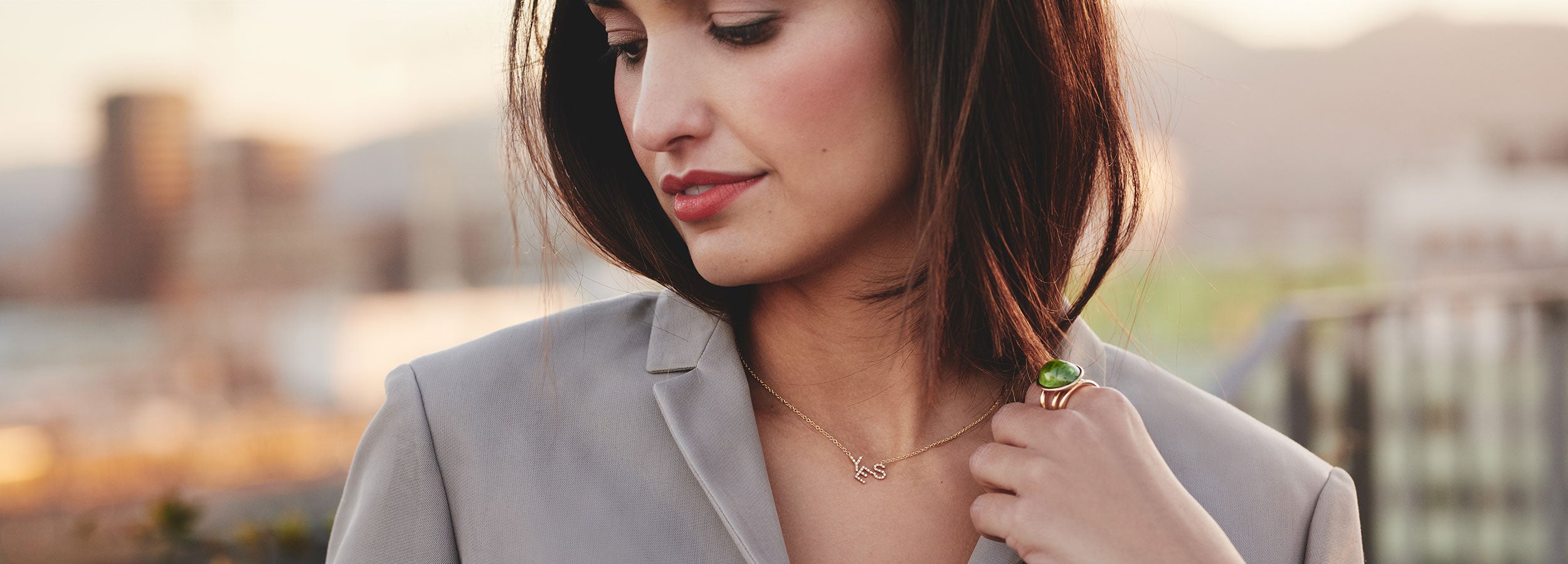 Eine Frau mit schulterlangem braunem Haar blickt nach unten und hält eine zarte Halskette in der Hand. Sie trägt einen grauen Blazer und einen großen grünen Ring. Der Hintergrund ist durch eine Außenkulisse sanft verschwommen.