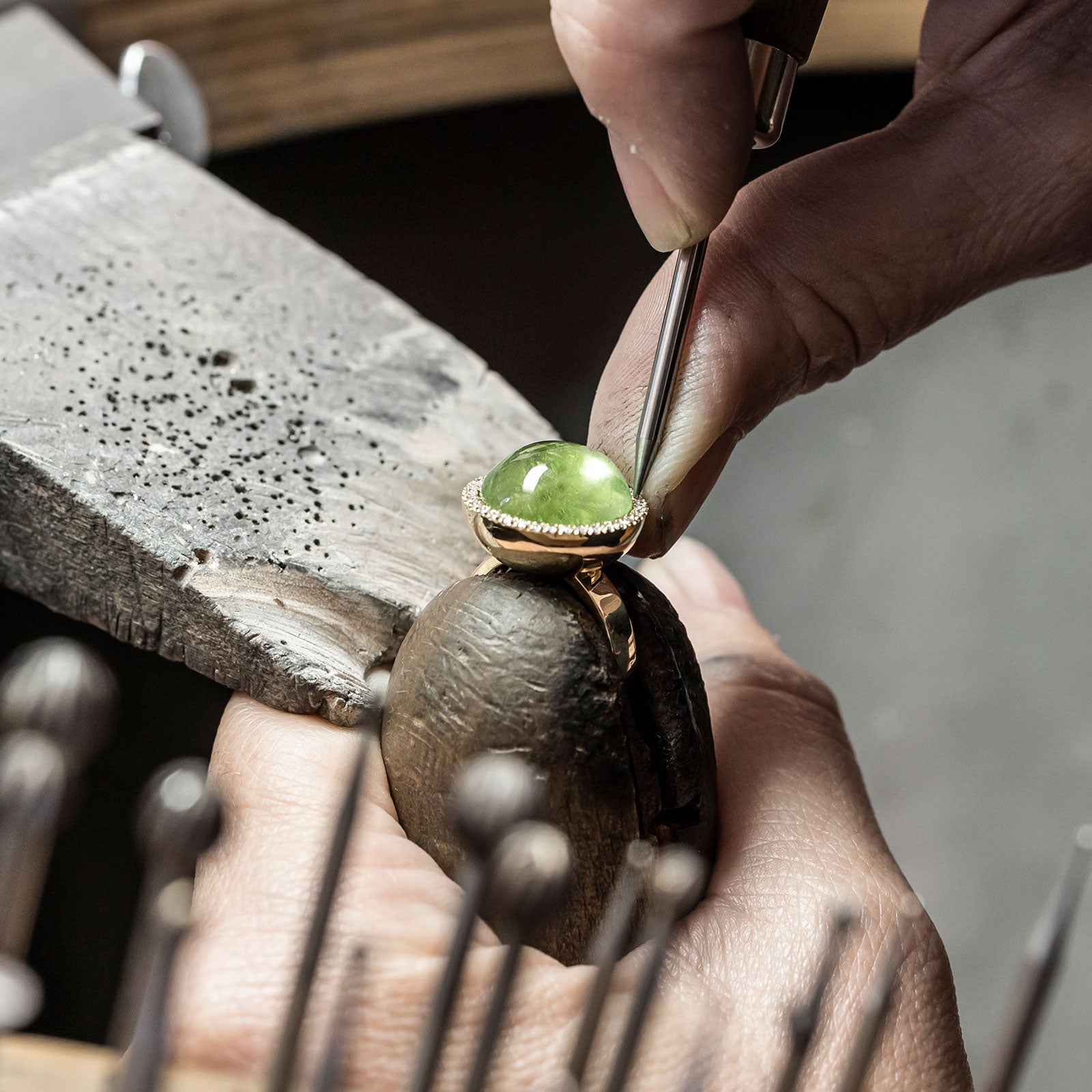 Ein Juwelier arbeitet an einem Ring, der einen grünen Edelstein hält, und verwendet ein Werkzeug zur Detaillierung. Der Ring ist auf einem Holzhalter befestigt. Im Vordergrund sind verschiedene Werkzeuge zu sehen, und eine strukturierte Metalloberfläche dient als Hintergrund.